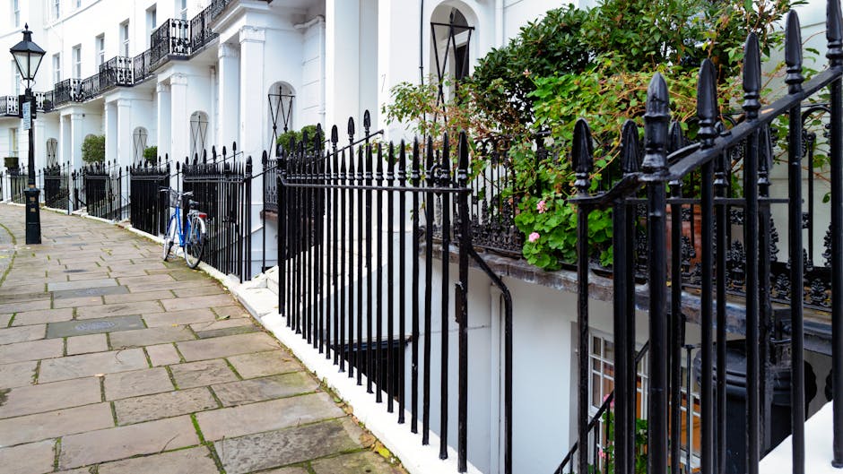A residential street featuring white terraced houses with black wrought iron railings bordering the sidewalk. The pavement is paved with large, rectangular stone slabs, some showing patches of moss an